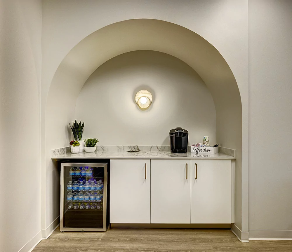 Small coffee station with a marble countertop, a Keurig coffee machine, potted plants, and a "Coffee Bar" sign, set within an arched alcove.